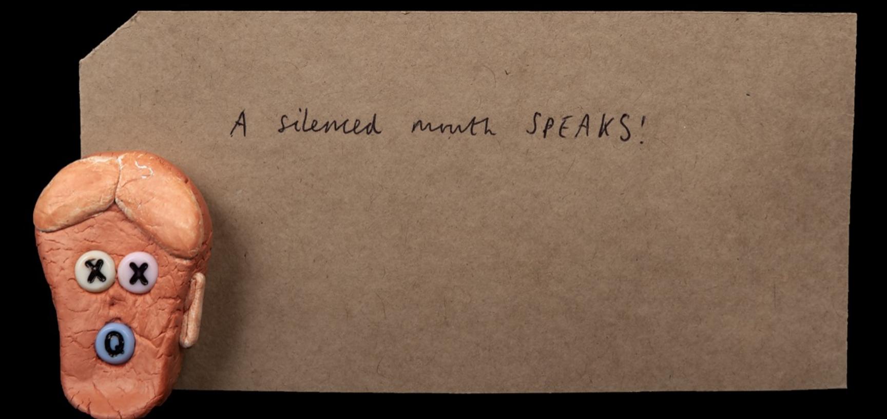 Very small clay head with crosses for eyes and an ‘O’ for a mouth. Sitting on top of a parcel tag reading ‘A silenced mouth speaks’.