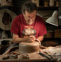 Portrait of Graham Taylor, wearing a read t-shirt, working with a large round piece of clay on a potters wheel, under the spotlight of a lamp